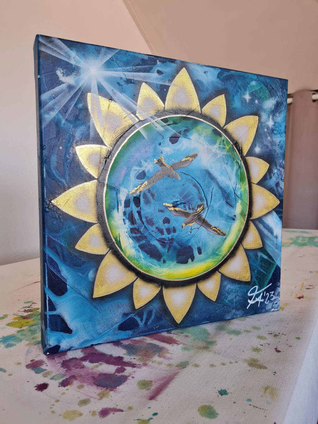 Gemaltes Bild mit einer blauen, gelb und grünen kreisförmigen Mitte, umgeben von goldfarbenen Blütenblättern, zwei goldgelb akzentuierten Vögeln fliegen in der Mitte, im Hintergrund Sterne und Leuchteffekte auf blauem Hintergrund.