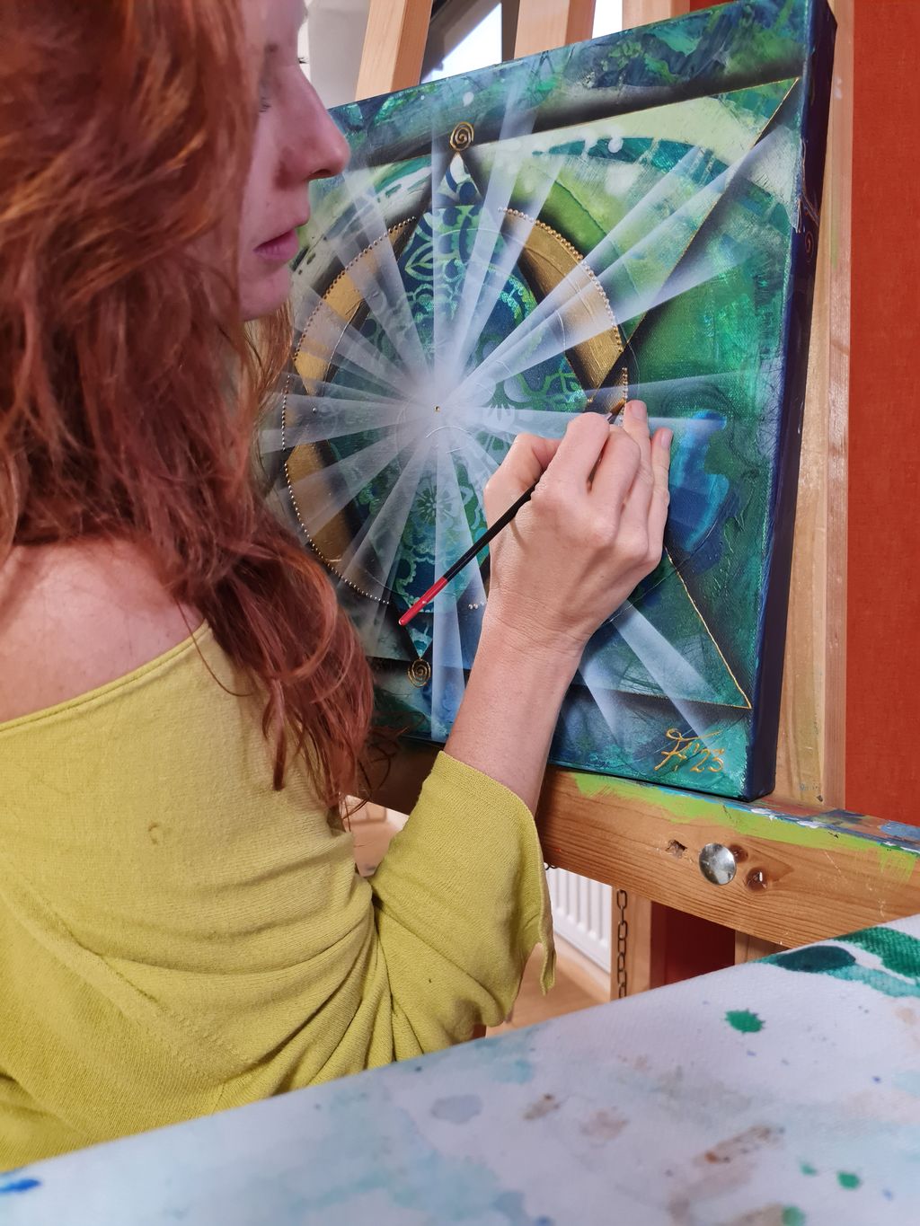 Frau mit roten Haaren trägt ein hellgrünes Oberteil und malt mit einem Pinsel ein geometrisches, farbenfrohes Bild auf eine Leinwand, die auf einer Staffelei steht.