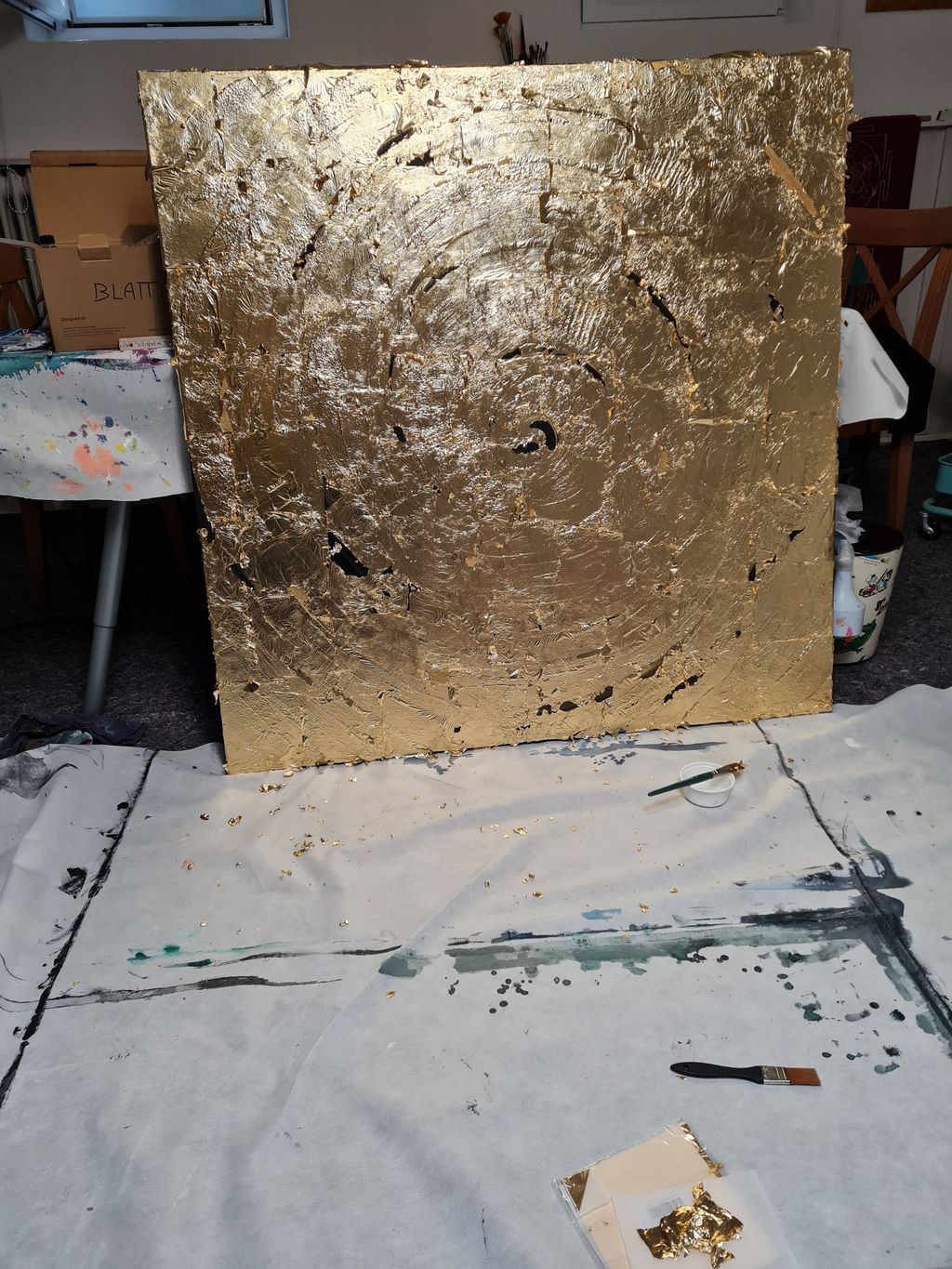 Eine in einem Raum stehende, großformatige Leinwand mit einer goldfarbenen, strukturierten Oberfläche, die kreisförmige Muster zeigt. Auf dem Boden davor liegen eine Malerbürste, ein Pinsel, ein kleines Gefäß und Goldfolie auf einer weißen Unterlage mit Farbspritzern.