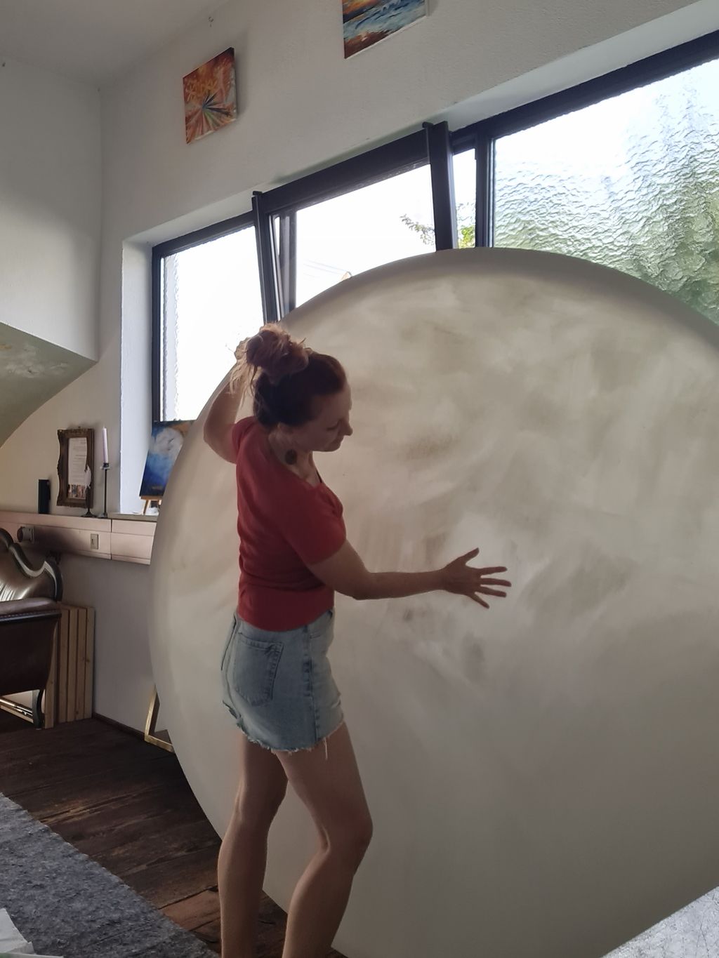 Eine Frau mit roten Haaren im Dutt trägt ein rotes T-Shirt und eine Jeans-Shorts und berührt spielerisch eine riesige runde, halbtransparente Leinwand in einem hellen Raum mit großen Fenstern.