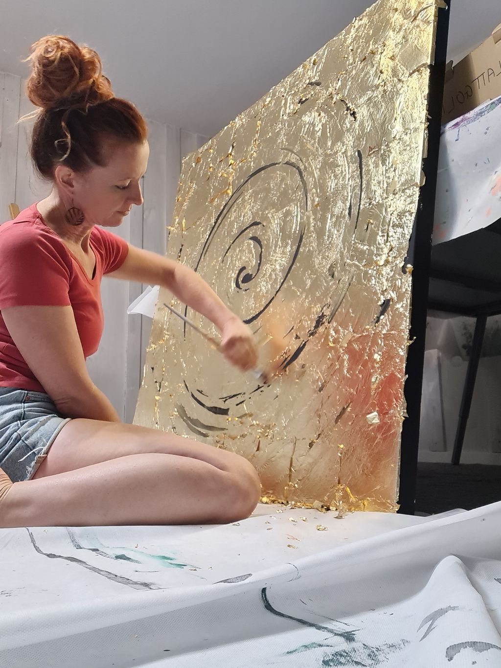 Frau mit roten Haaren sitzt auf dem Boden und arbeitet mit einem Pinsel an einem großen goldenen Kunstwerk mit spiralförmigem Muster.