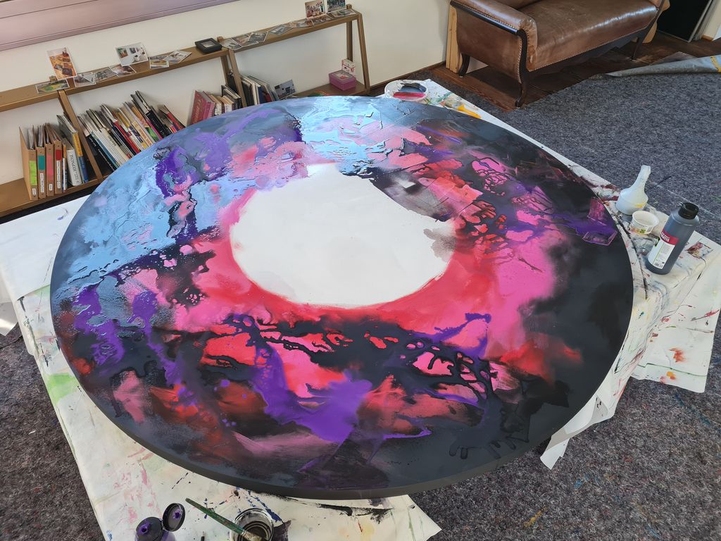 Großes rundes Leinwandbild mit abstrakten Mustern in Schwarz, Rot, Violett und Blau auf einem Tisch in einem Raum mit Teppichboden, Bücherregalen und einem Ledersofa.