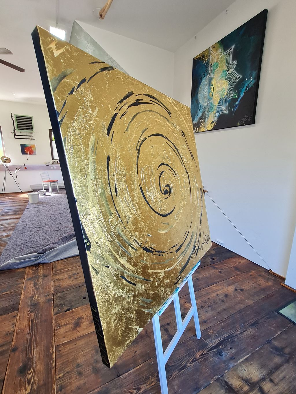 Leinwand mit einem goldenen, spiralförmigen abstrakten Muster auf einer Staffelei in einem Raum mit Holzboden und weiß gestrichenen Wänden. Im Hintergrund hängt ein weiteres farbenfrohes Kunstwerk an der Wand.