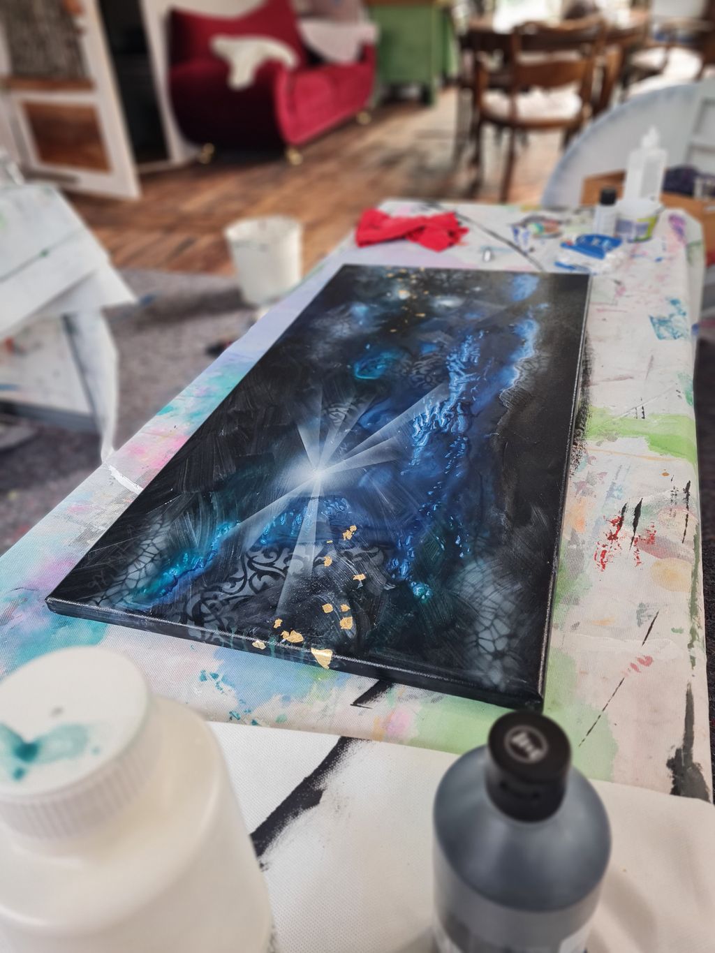 Ein abstraktes Bild mit einem leuchtenden weißen Lichtstern in der Mitte und blauen sowie schwarzen Farbtönen, liegt auf einem bemalten Tisch in einem Atelier mit Holzfußboden und rotem Sofa im Hintergrund.