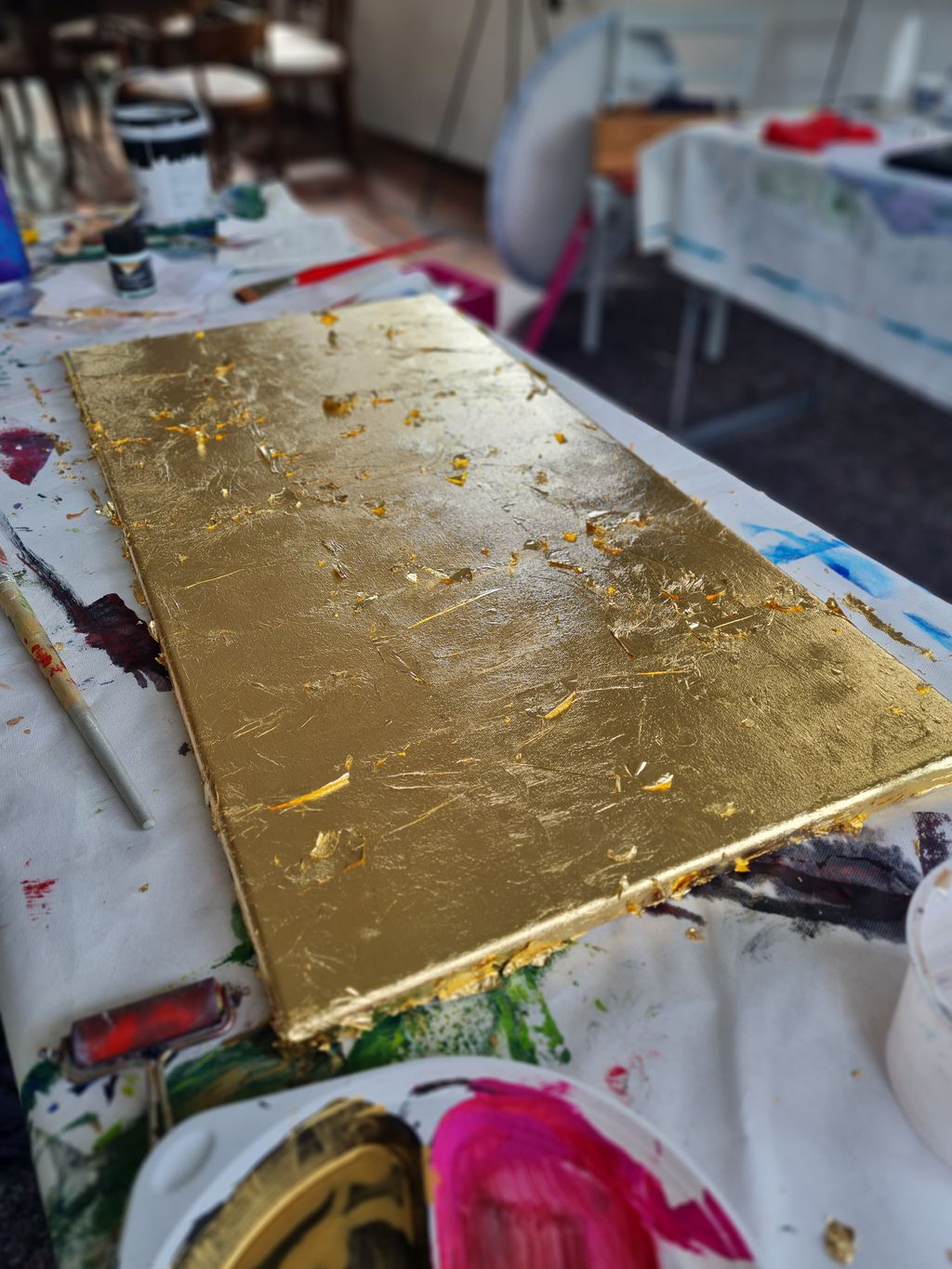 Ein rechteckiges Werkstück, das mit unregelmäßigem Blattgold auf einer bemalten Arbeitsfläche bedeckt ist, umgeben von Farbeimern und Pinseln in einem Atelier.