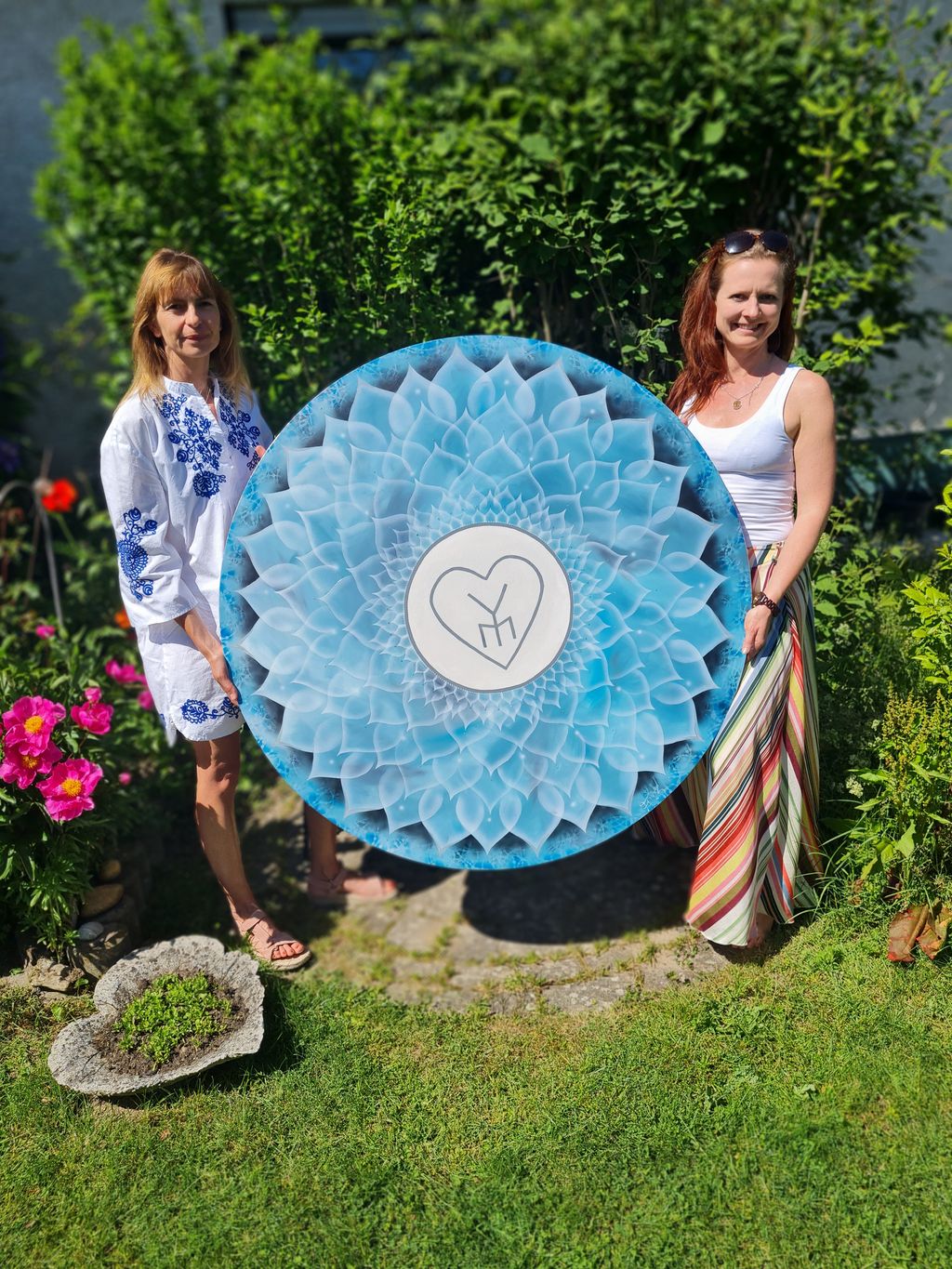 Zwei Frauen im Garten halten gemeinsam eine große, runde, blau-weiß gemusterte Scheibe mit einem Herzsymbol in der Mitte.