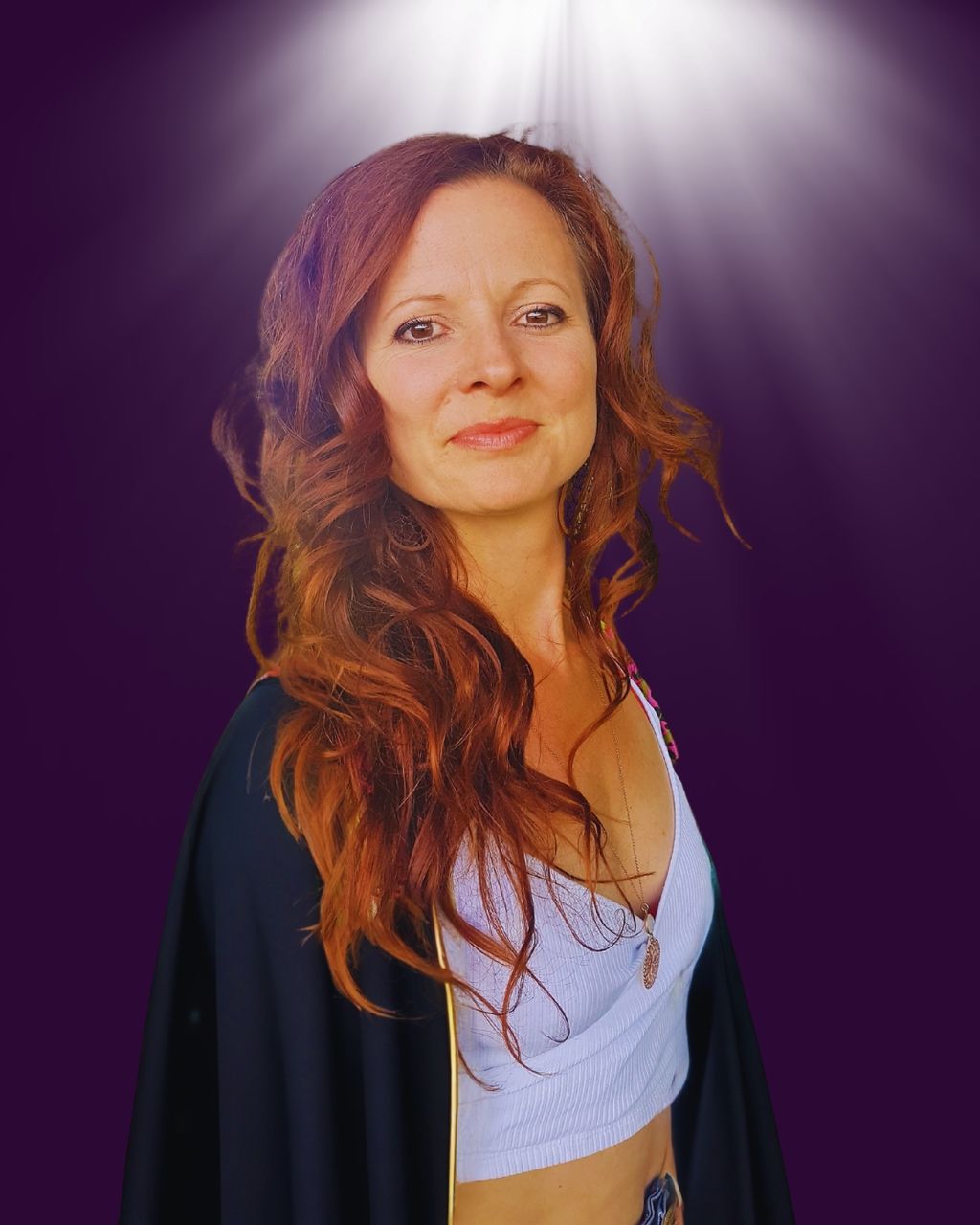 Frau mit langen, roten lockigen Haaren trägt ein weißes Crop-Top und einen schwarzen Umhang vor einem violetten Hintergrund mit Lichtstrahlen von oben.