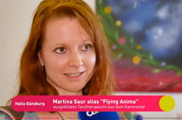 Porträt einer rothaarigen Frau, die in einem Interview spricht; im Hintergrund unscharfes, farbenfrohes Wandbild. Ein Schriftzug zeigt ihren Namen und Beruf als ausgebildete Tanztherapeutin.