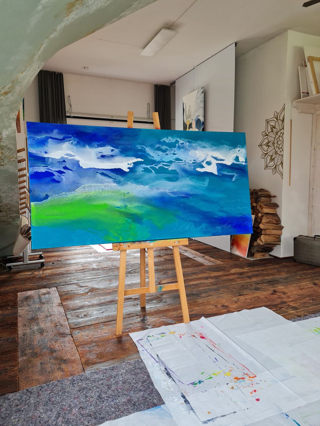 Ein abstraktes Gemälde mit blauen und grünen Farbtönen auf einer Staffelei in einem Künstlerstudio mit Holzboden und weißen Wänden.