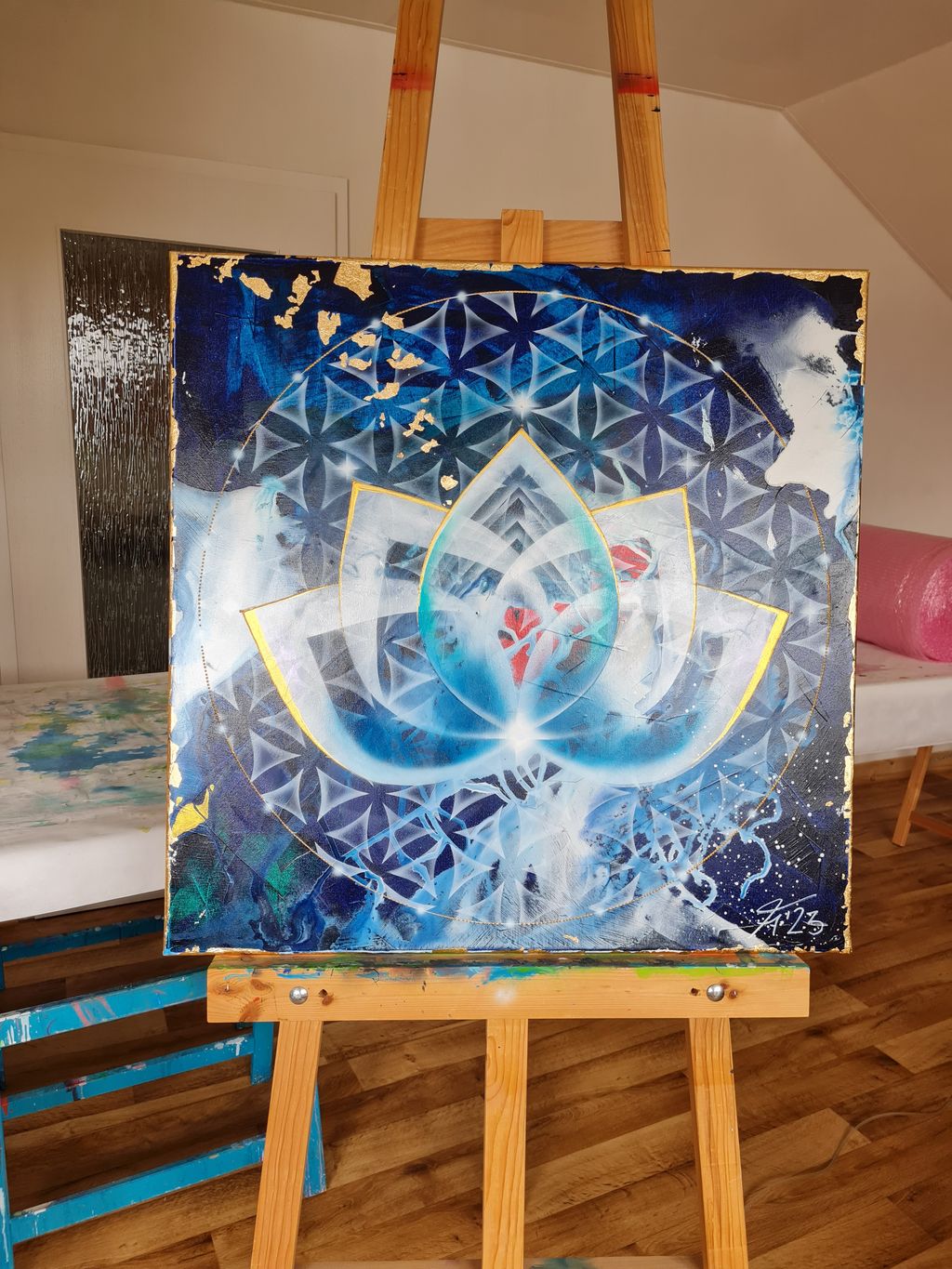 Ein auf einer Holzstaffelei stehendes Gemälde in einem Raum mit Holzfußboden, das eine abstrakte, geometrische Lotusblume in Blau- und Weißtönen mit goldenen Akzenten zeigt.