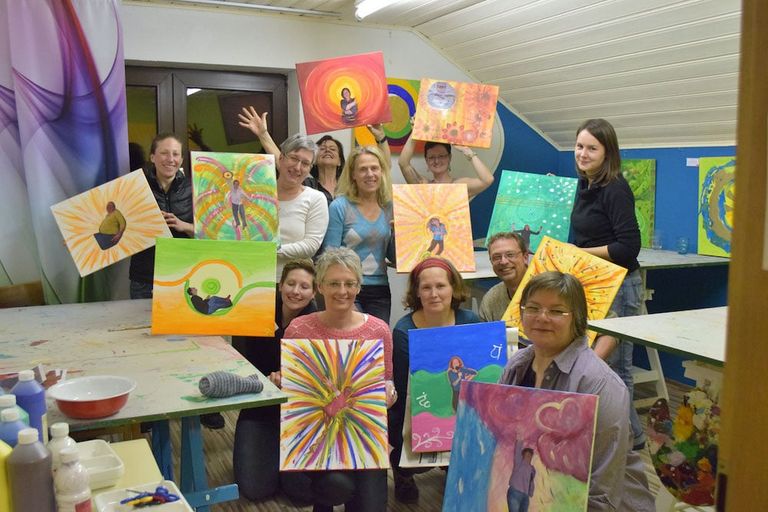 Eine Gruppe von zehn Personen in einem Atelier, die jeweils farbenfrohe Gemälde mit abstrakten und figurativen Motiven präsentieren und lächeln.