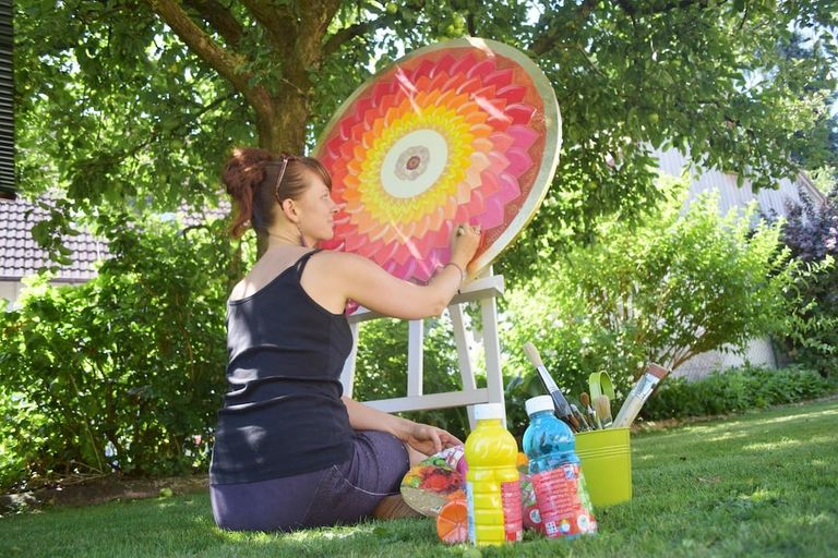 Frau sitzt auf dem Gras in einem Garten und malt ein buntes, kreisförmiges Mandala auf eine Leinwand, mit verschiedenen Farben und Pinseln neben ihr.