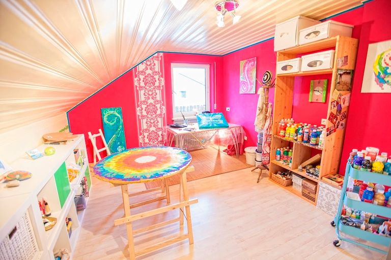Atelier unter einer schrägen Decke mit roten Wänden, einem runden, farbenfrohen Tischtuch auf einem Holztisch und Regalen mit vielen Farbflaschen und Kunstwerken an den Wänden.