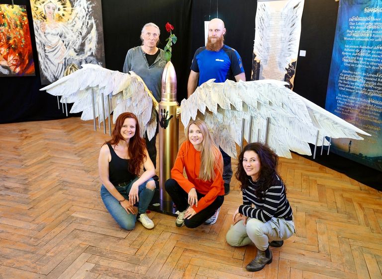 Fünf Personen posieren in einer Kunstgalerie um eine Skulptur mit zwei großen, weißen Flügeln und einer goldfarbenen Kugel mit einer roten Rose oben drauf.