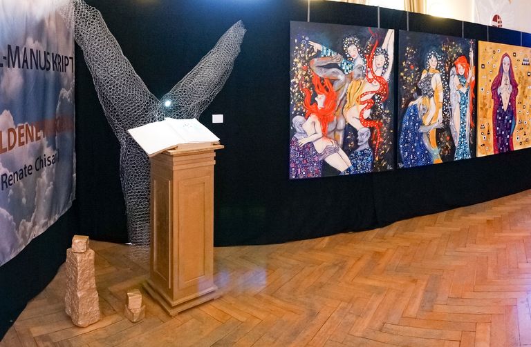 Ausstellung mit Gemälden von Frauen in bunten Kleidern an einer schwarzen Wand und einem Podest mit einem offenen Buch, umgeben von Drahtskulptur und Steinfiguren, auf einem Parkettboden.
