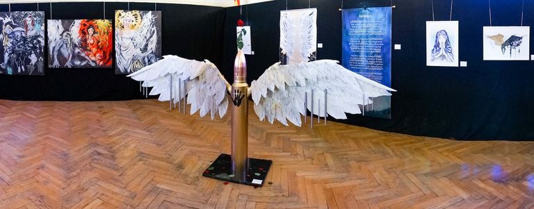 Skulptur einer großen Kugel mit weißen Flügeln in einem Ausstellungsraum mit Holzfußboden und mehreren Kunstwerken an den Wänden.