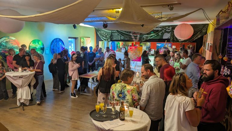 Menschen stehen und unterhalten sich in einem farbenfroh dekorierten Raum bei einer Ausstellung oder Veranstaltung mit Kunstwerken an den Wänden und Stehtischen.