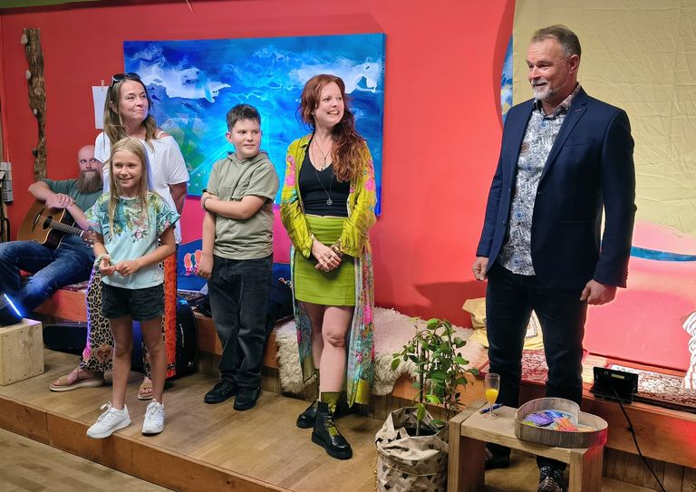 Fünf Personen stehen und sitzen in einem farbenfrohen Raum mit roter Wand und blauem Gemälde, ein Mann spielt Gitarre, während die anderen lächeln und sich unterhalten.