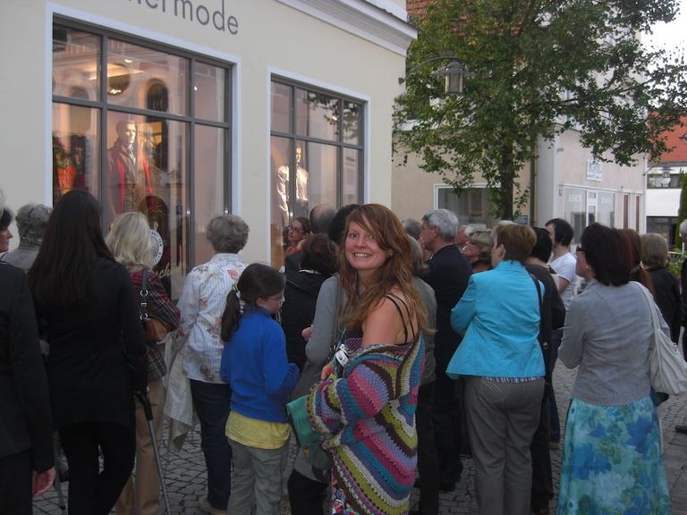 Menschenmenge vor einem Schaufenster mit Männerfiguren und Ausstellungsbeschriftung an einem Gebäude in einer gepflasterten Straße mit Bäumen.