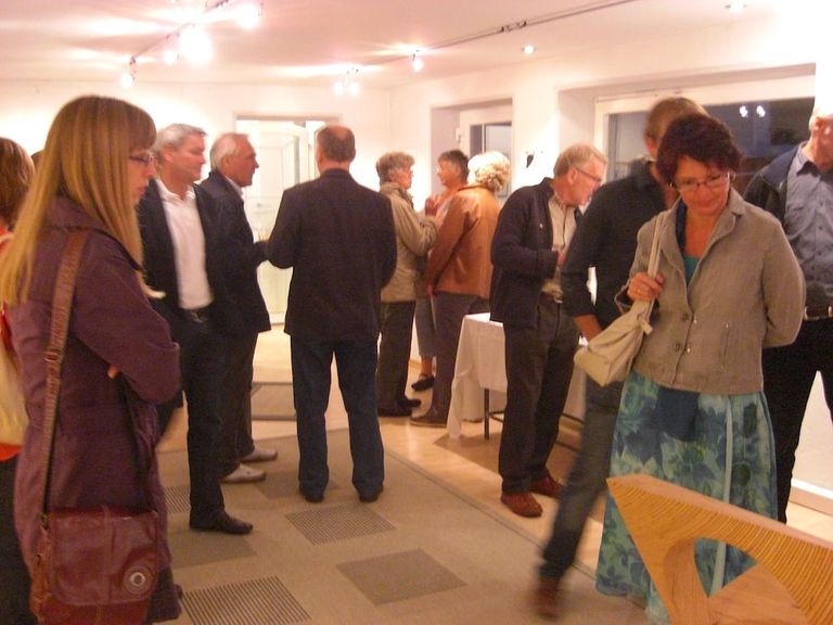 Mehrere Personen unterhalten sich und betrachten Kunstwerke in einer gut beleuchteten Ausstellungsraum mit weißen Wänden.