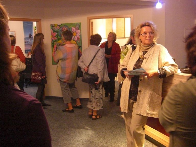Mehrere Frauen betrachten und diskutieren Kunstwerke in einer Innenraumausstellung, während eine Frau mit Brille und beige Jacke Papiere hält und an einem Holzpult lehnt.