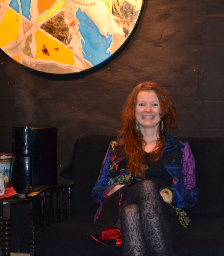 Eine rothaarige Frau mit farbenfrohem Jackett und schwarzem Kleid sitzt lächelnd auf einem dunklen Sofa vor einer schwarzen Wand mit einer bunten, runden Wanddekoration.