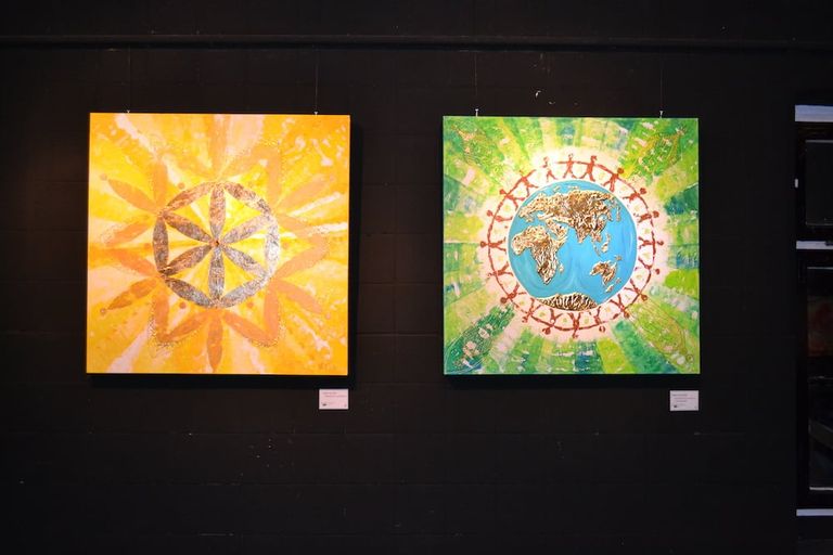 Zwei quadratische Gemälde an einer schwarzen Wand: Links ein gelbes Bild mit einem geometrischen Muster, rechts ein grünes Bild mit der Weltkarte in der Mitte, umgeben von roten menschlichen Figuren, die Hände halten.