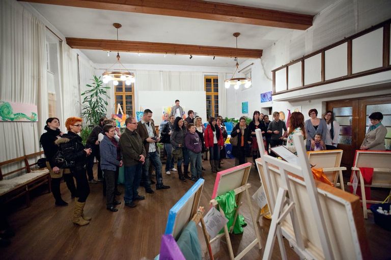 Gruppe von Menschen, die in einem großen, gut beleuchteten Raum an einer Kunstausstellung mit leeren Leinwänden auf Staffeleien teilnehmen.