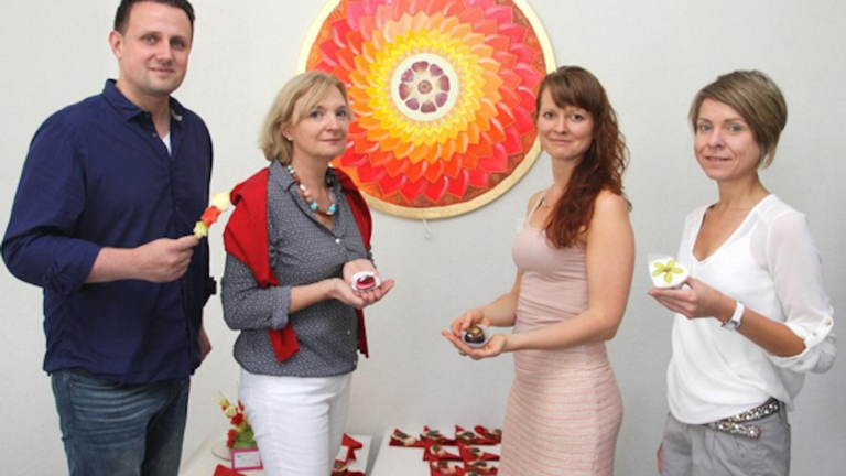 Vier Personen stehen vor einer Wand mit einem großen, runden Kunstwerk mit roten, gelben und orangen Farbtönen und halten kleine kunstvoll dekorierte Speisen oder Desserts in den Händen.