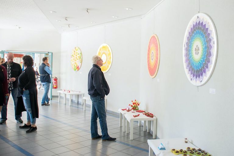 Mehrere Menschen betrachten bunte, runde Kunstwerke an der Wand in einem hellen Ausstellungsraum mit Fliesenboden.