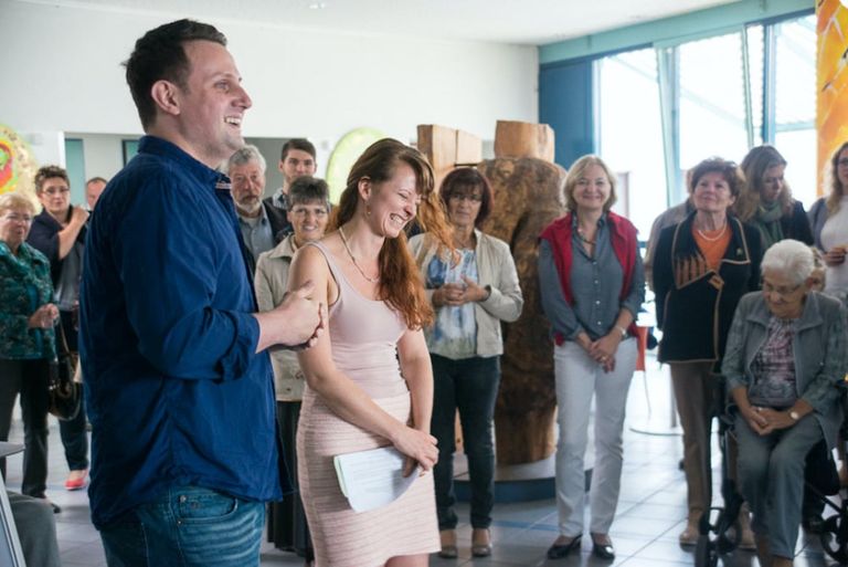 Eine Gruppe von Menschen steht in einem hellen Raum, zwei Personen vorne lachen und halten Papiere, während die anderen im Hintergrund aufmerksam zuhören.