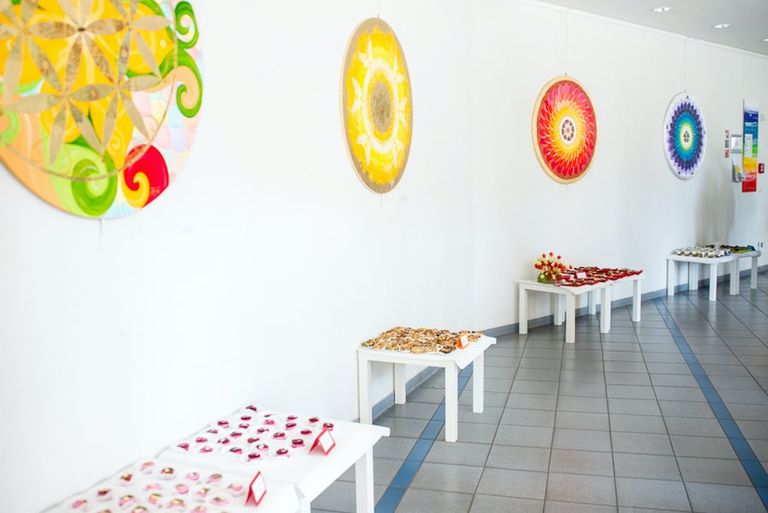 Eine Ausstellungsfläche mit weißen Tischen, auf denen verschiedene Schmuckstücke liegen, darunter Ringe und Armbänder, an einer weißen Wand hängen bunte, runde Mandala-Kunstwerke.