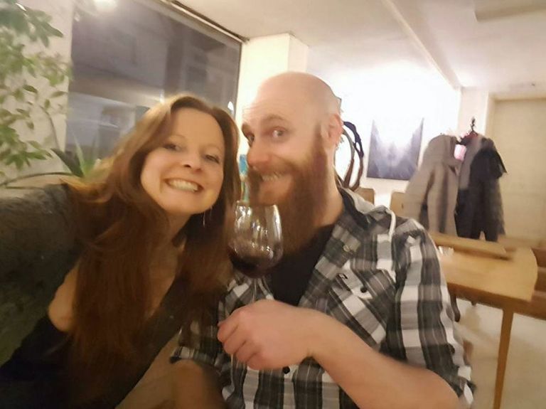 Zwei lächelnde Personen in einem Innenraum, eine hält ein Glas Rotwein, im Hintergrund Garderobe und Kunstwerke an der Wand.