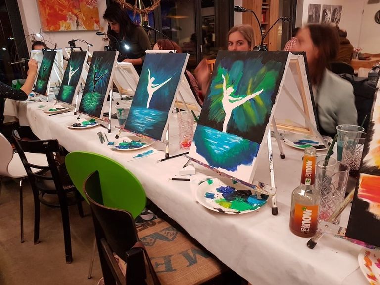 Mehrere Staffeleien mit Gemälden eines tanzenden Balletttänzers vor einem bunten Hintergrund auf einem langen Tisch, an dem mehrere Personen teilnehmen, einige mit Pinsel in der Hand.
