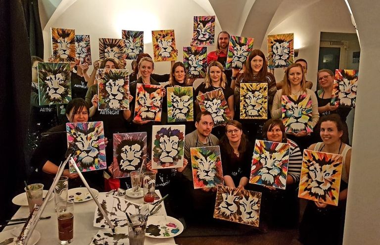 Gruppe von Menschen in einem Raum, die jeweils ein gemaltes Bild eines stilisierten Löwenkopfs in verschiedenen Farben hochhalten, umgeben von Malutensilien auf Tischen.