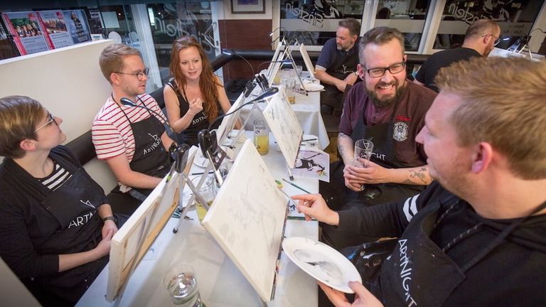 Mehrere Personen sitzen an Tischen in einem Atelier und malen auf Staffeleien, während sie miteinander sprechen und Getränke genießen.