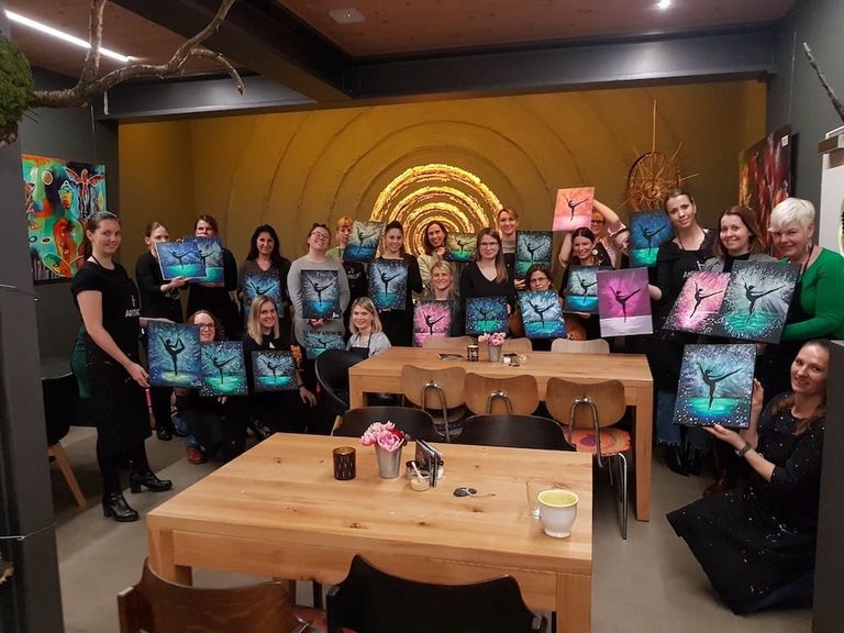 Eine Gruppe von Frauen in einem Raum mit gelber Wand und bunten Kunstwerken, die eigene Bilder mit einer Tänzerin vor farbigen Hintergründen halten.