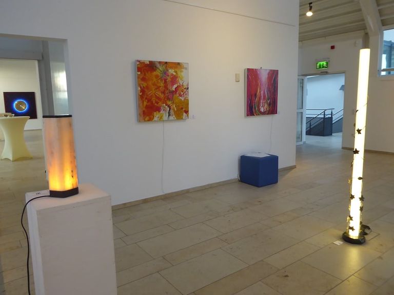 Innenraum einer Galerie mit bunten Gemälden an der weißen Wand und zwei beleuchteten Stehlampen, eine links auf einem weißen Podest und eine rechts mit Pflanzenmotiven.