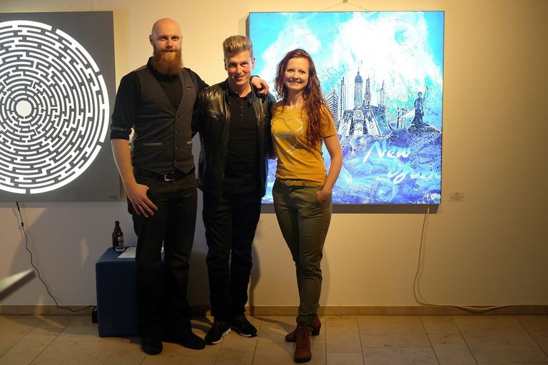 Drei Personen stehen vor zwei modernen Kunstwerken auf einer Ausstellung, eine beleuchtete labyrinthartige Grafik und ein blaues Gemälde mit Skylines und Schriftzug.