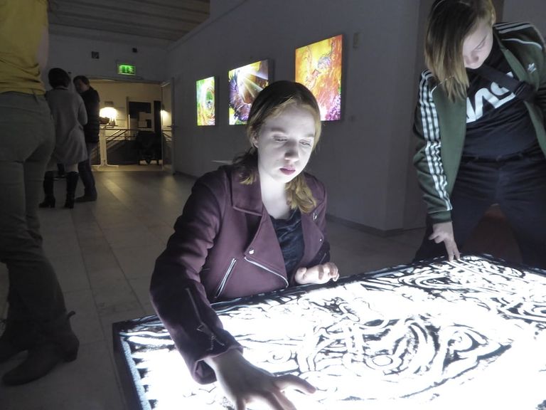 Zwei Jugendliche interagieren mit einem beleuchteten Tisch mit abstrakten Mustern in einer Galerie mit Kunstwerken an der Wand.