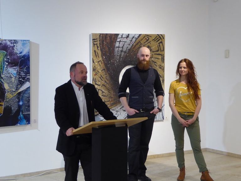 Drei Personen in einer Galerie mit moderner Kunst an den Wänden, von denen eine an einem Rednerpult steht und spricht.