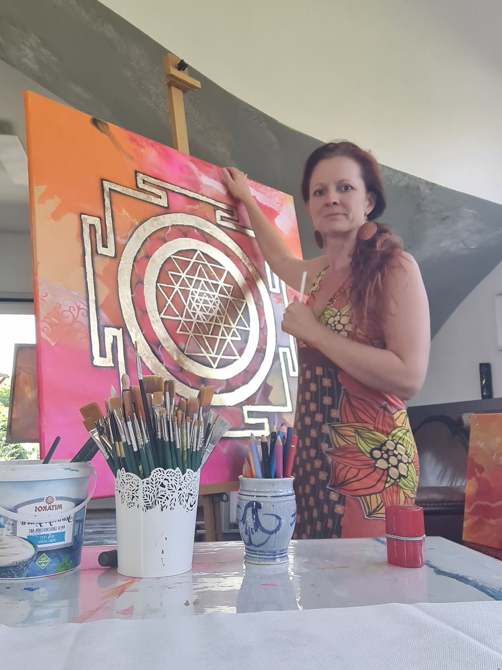 Frau in bunter Kleidung steht vor einer Staffelei und malt ein großes, goldfarbenes Sri-Yantra-Mandala auf orange-rosa Hintergrund.