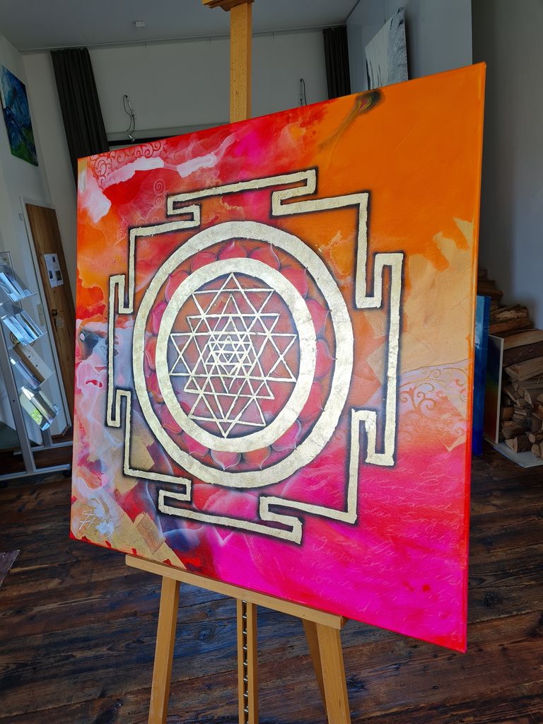 Ein farbenfrohes Gemälde auf einer Staffelei in einem Raum, das ein goldenes Sri Yantra vor einem Hintergrund aus Orange, Rosa und Rot zeigt.