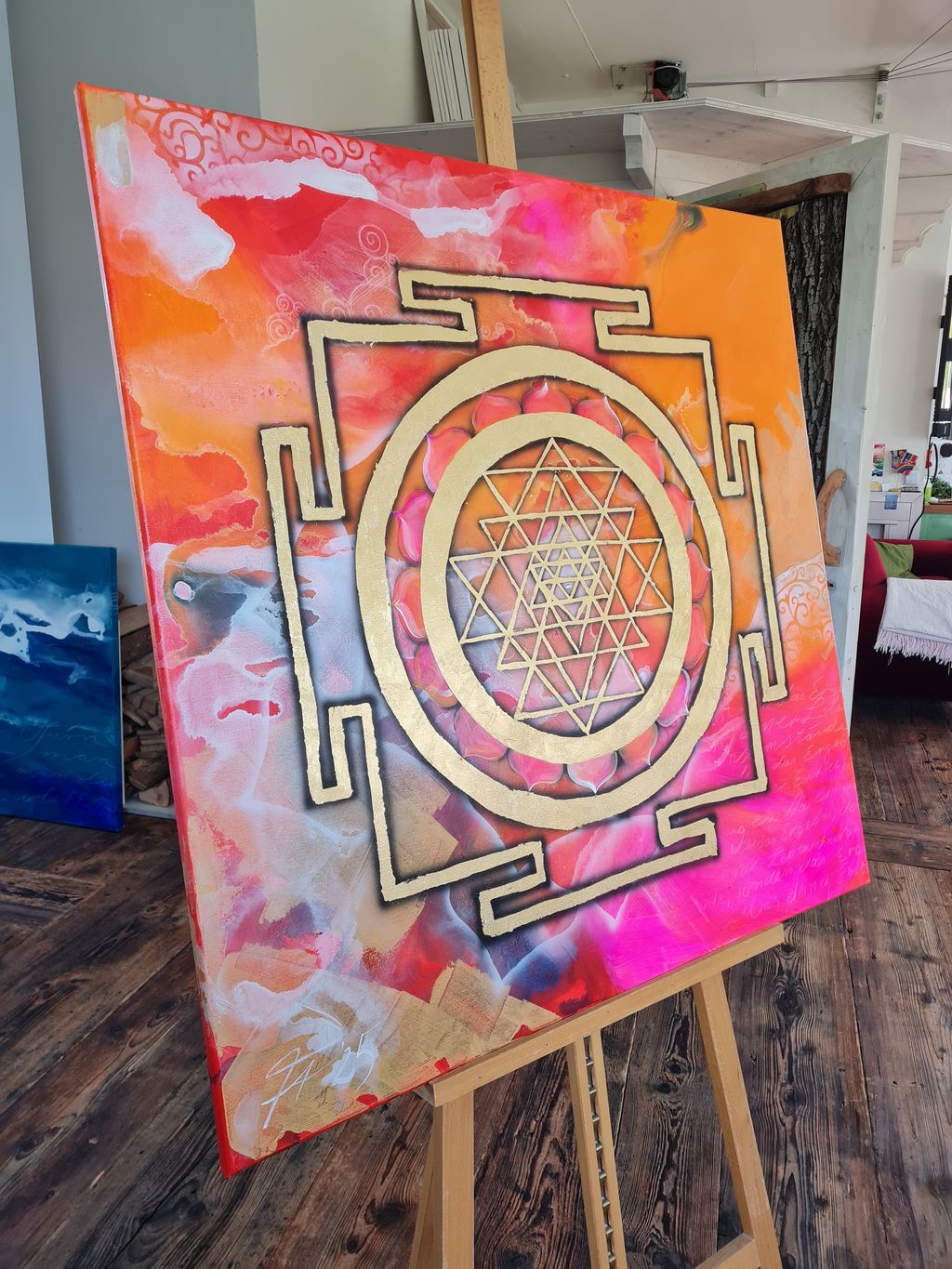 Leinwand mit einem leuchtend orange-rosa Hintergrund, darauf ein goldenes Sri Yantra-Symbol, das auf einem hölzernen Staffelei steht. Im Hintergrund ein Raum mit Holzboden, Möbeln und weiteren Kunstwerken.