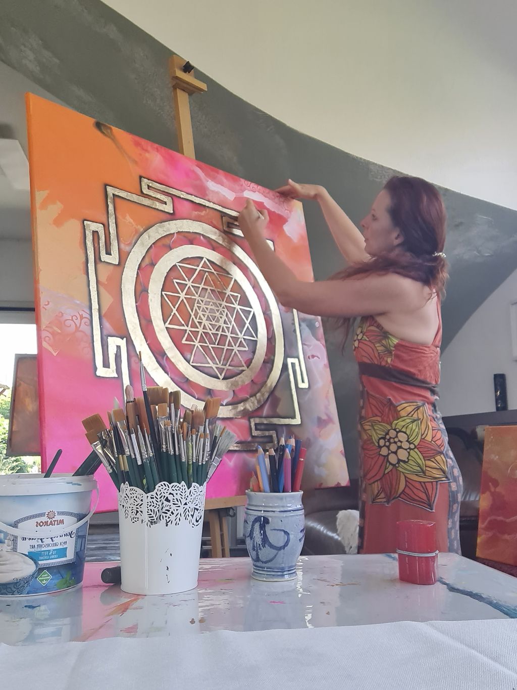 Frau malt ein großes orange-pinkes Leinwandbild mit goldenem Sri Yantra Symbol in einem gut beleuchteten Raum.