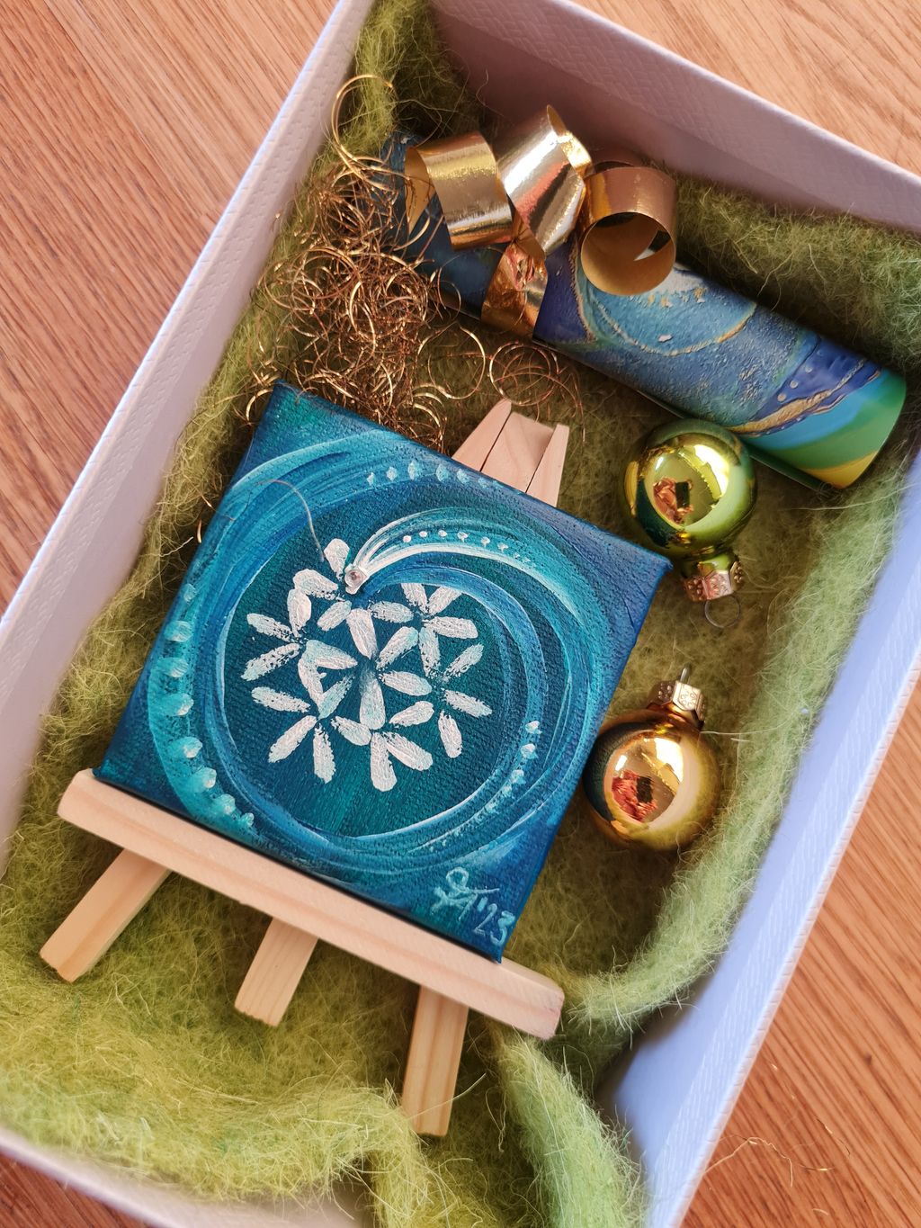Eine Geschenkbox mit grüner Einlage enthält eine kleine Staffelei mit einem blau gemalten, weißen Mandala-Muster, zwei goldene Weihnachtskugeln, goldene Deko-Spiralen und eine mit blau-grünem Muster verzierte Rolle.