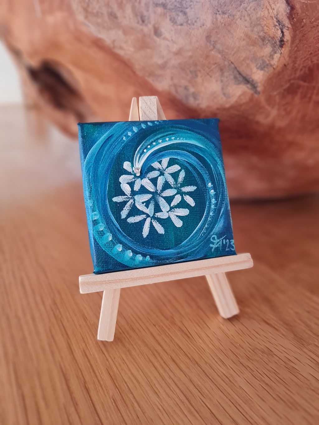 Kleine blau bemalte Leinwand mit abstraktem weißen Blumenmuster auf einem Mini-Holzstaffelei stehend auf einer Holzoberfläche.
