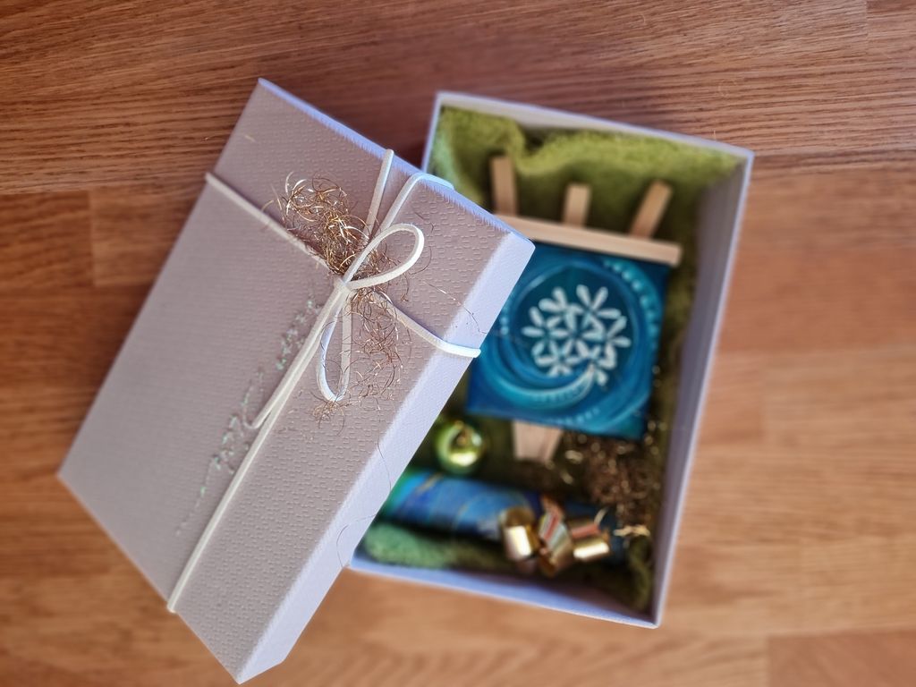 Offene Geschenkbox mit grauem Deckel und weißem Band, darin ein kleines Holzstaffelei mit einem blauen Kunstwerk, eine dekorative goldene Kugel und goldfarbene Geschenkbandstücke auf grünem Stoff.