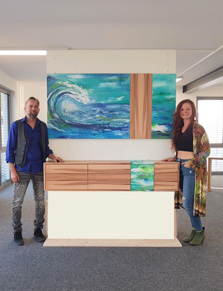 Ein Mann und eine Frau stehen jeweils links und rechts neben einem Holzsideboard mit einem blauen und grünen Gemälde im Hintergrund, das eine große Welle im Meer zeigt, in einem modernen Innenraum.
