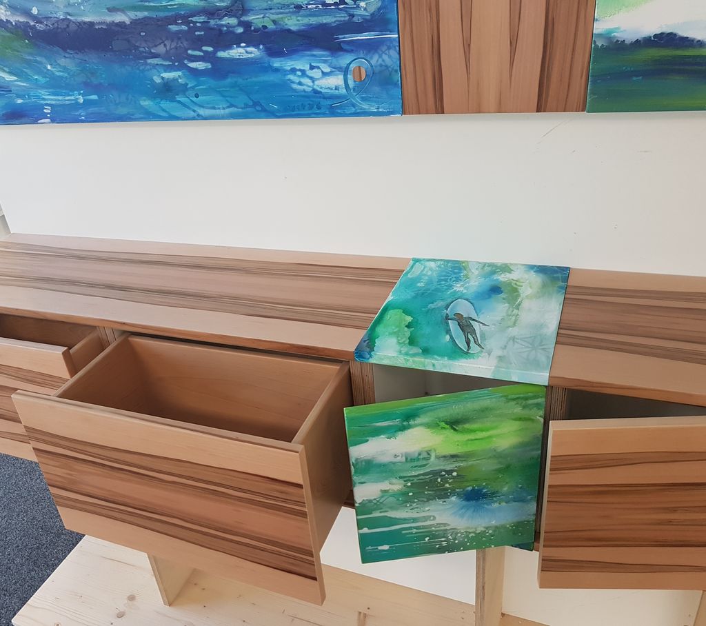 Offene hölzerne Schubladen mit künstlerisch bemalten Fronten in Blau- und Grüntönen, die abstrakte und naturinspirierte Muster zeigen, in einem modernen Raum mit Gemälden an der Wand.